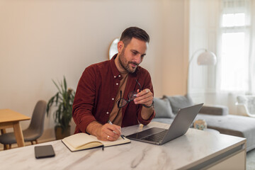 A hard-working young adult man working from home on his laptop while taking notes in a notebook