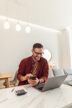 A smiling adult male accountant using his phone and a laptop while working from home