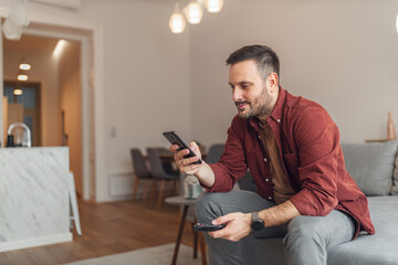 A happy adult man scrolling through his phone while relaxing on a couch in the living room