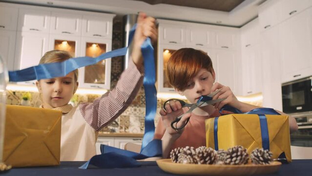 Slow motion shot footage of brother and sister sitting at table in kitchen preparing gift boxes for Christmas together