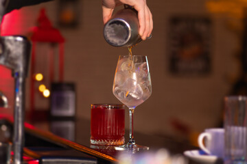 Close-up of the bartender carefully pours a ready-made cocktail from a metal shaker into a glass with ice behind the bar.