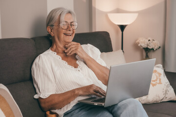 Happy amused senior white haired woman sitting on home sofa watching a funny movie on laptop enjoying tech and social