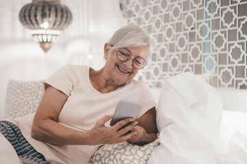 Smiling senior woman at bedroom at home or hotel room looking at smartphone enjoying tech and social. Elderly female online with cellphone