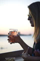 giovane donna beve un cocktail al tramonto in riva al mare - 772994841