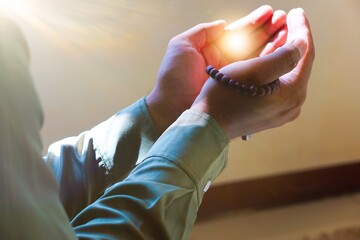 the hands of a man in Muslim prayer holding prayer beads, with light between his palms