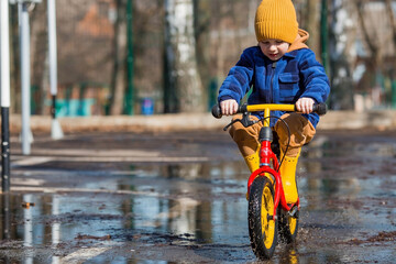 A cheerful little boy rides a bike through puddles. A happy child walks outside in the spring. The...