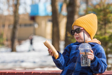 A little boy eats fast food outside in the spring. He will rub the baby in a fashionable plaid shirt, yellow hat and sunglasses. The child eats a hot dog and drinks lemonade. - obrazy, fototapety, plakaty