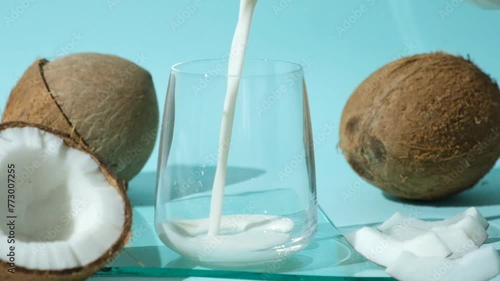 Wall mural Coconut milk pouring into a glass among coconuts on a blue background, slow motion.