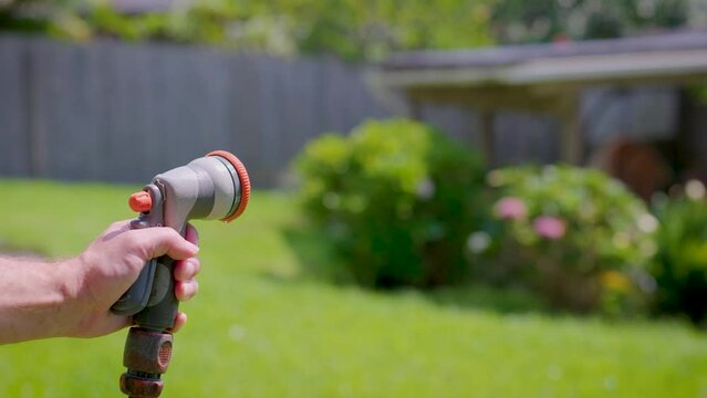 Spraying water with garden hose and adjustable nozzle in slow motion. Man's hand holding spray gun and watering plants, grass in backyard.