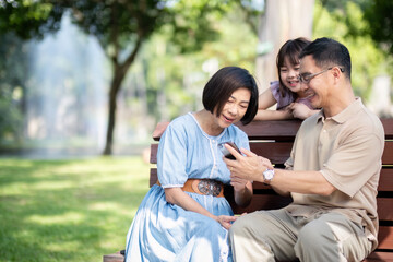 Family, generation and technology concept happy smiling grandmother, grandfather and little granddaughter with telephone sitting at park
