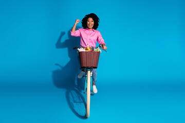Full length photo of overjoyed funky girl dressed silk shirt jeans riding bike raising up fist win bet isolated on blue color background