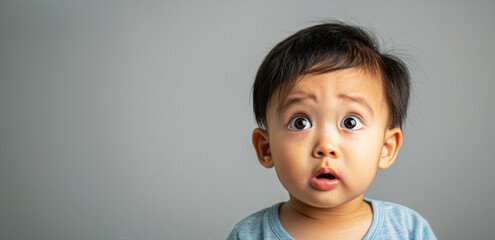 A baby with a surprised expression on his face. The baby is wearing a blue shirt. The image has a gray background. a 1-year-old Asian toddler, bewildered expression, looking confused and dazed - obrazy, fototapety, plakaty