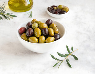 A bowl of olives is on a table next to a bottle of olive oil. The olives are green, black, and purple, and they are arranged in a way that makes them look like a small salad