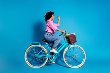 Full length side profile photo of excited girl dressed shirt riding bike look empty space clenching fist isolated on blue color background