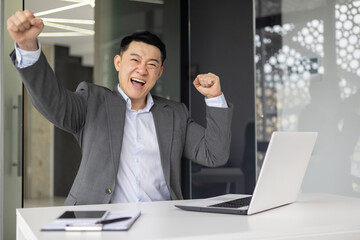 Triumphant Asian businessman at his desk in an office raising his fist in victory or success while working on his laptop.