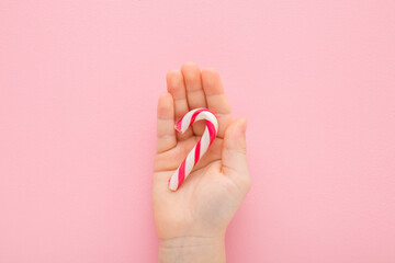 Red white candy cane in little baby girl opened palm on light pink table background. Pastel color. Children sweet snack. Closeup. Top down view.