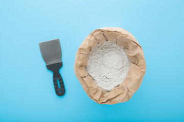 Spatula and opened paper bag of white dry finishing putty for plastering ceiling and wall surfaces on light blue table background. Pastel color. Repair work of home. Closeup. Top down view.