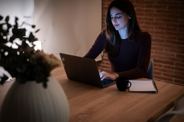 Concentrated young woman working late in home office, using laptop, copy space. Casual girl in the light of the screen on the background of the illuminated room. Technology and work concept