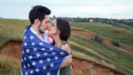 Happy couple of man and woman kiss wrapped in American flag on hill against village houses. Patriotism and romantic relationships, close-up