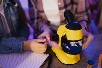 Close up of hands of mother controlling process of creating of microscope slide for study of plant cells by her little daughter. Young woman teaching girl to make biological research at evening home.