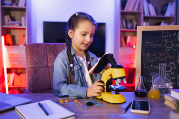 Focused little girl in denim shirt learning science in evening at home using electron microscope and smartphone. Concept childhood and studying at school.