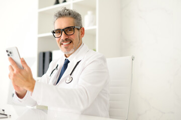 A senior doctor with grey hair is pictured in a well-lit clinic, holding a smartphone, staying...
