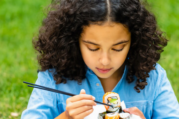 girl in nature. little tanned girl with dark curly hair sits on green grass and eats sushi with chopsticks, food concept