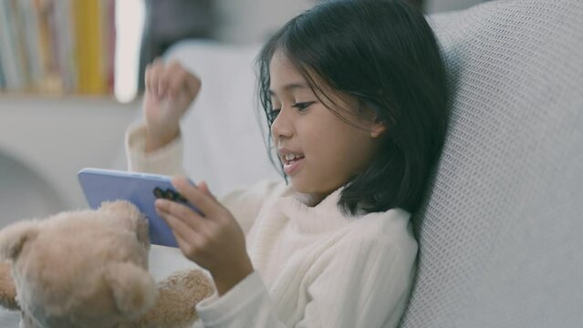 A young girl is sitting on a couch with a teddy bear and playing a video game
