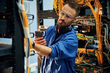 Holding smartphone. Young man is working with internet equipment and wires in server room