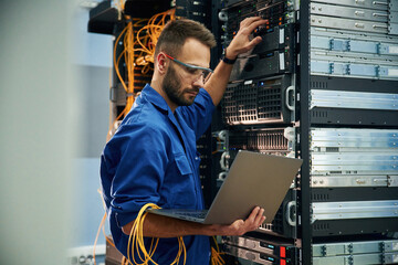 Using the laptop. Young man is working with internet equipment and wires in server room