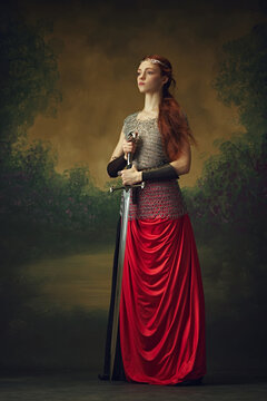 Portrait of knight-queen with flowing red hair, clad in armor and crimson, standing firm with her blade against vintage background. Concept of historical and modern times, beauty and fashion. Ad