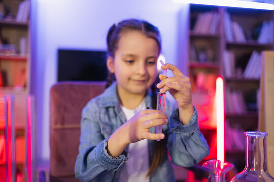 Focused Caucasian little girl doing chemistry experiment looking at test tube in evening at living room. Science, hobbies, learning, education, discovery, childhood and domestic life.