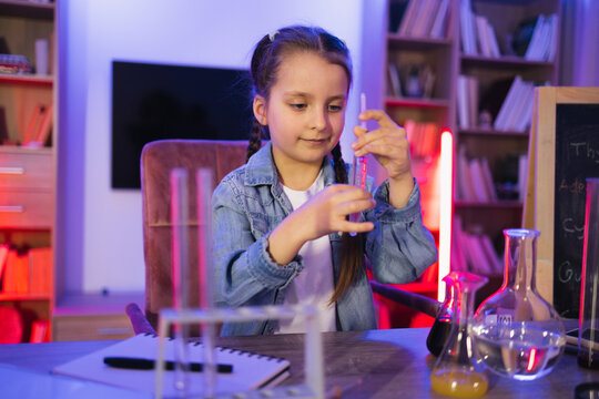 Focused Caucasian little girl doing chemistry experiment looking at test tube in evening at living room. Science, hobbies, learning, education, discovery, childhood and domestic life.