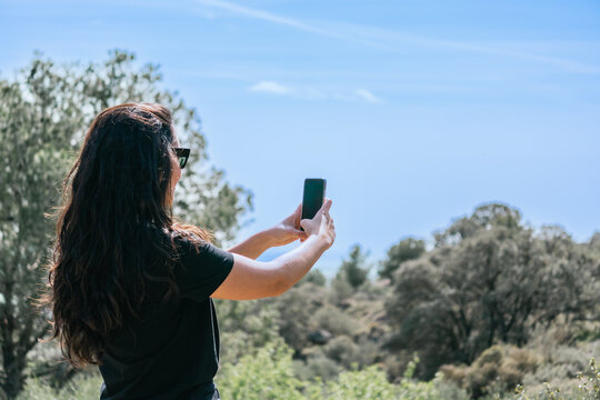 A woman is taking a picture of the landscape with her cell phone, she's on a natural environment