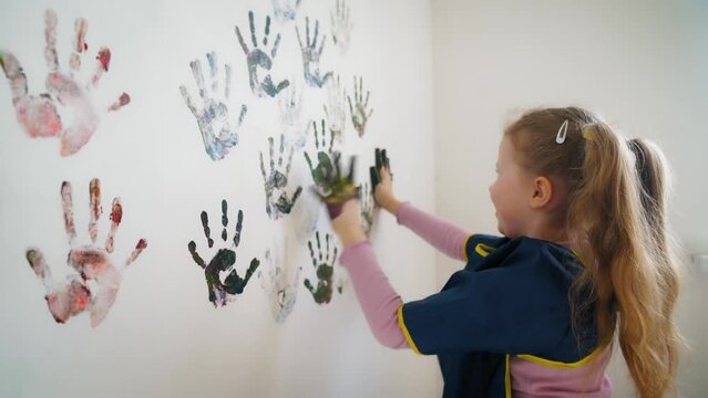 Six year old cheerful girl leaves colored handprints on white wall wall in room. Children's palms dipped in paint make prints on the wall. 4k footage