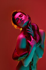 Beautiful young shirtless woman with slim body posing with snake against red studio background in neon light. Sensuality and tenderness. Concept of female beauty, animal theme, care, elegance, fashion
