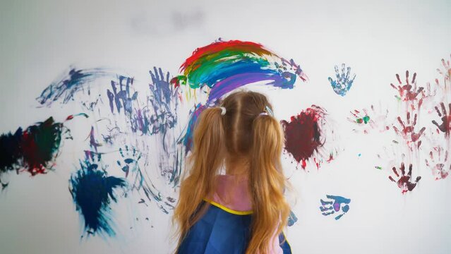 Mischievous cheerful girl child paints with multicolored paints white wall wall in the room. View from the back, daughter drawing a rainbow on the wall. 4k footage.