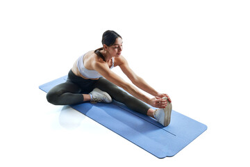 Fototapeta premium Young athletic woman in white tank-top and legging stretching legs sitting on mat against white studio background. Concept of sport and recreation, movement, self care, action, energy. Ad