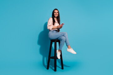 Full length photo of pretty shiny lady dressed beige shirt sitting chair texting modern gadget isolated blue color background