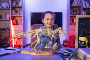 Foto op Canvas Portrait of smiling little girl sitting at table and examine skeleton of dinosaur. Caucasian child in casual wear making model of tyrannosaurus out of bones in evening at home. © sofiko14