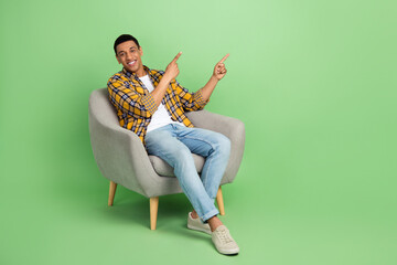 Full length portrait of positive guy sit cozy chair indicate fingers empty space offer isolated on...