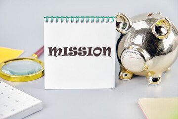 Concept of mission word on a white notebook on a gray background near a piggy bank, a calculator and stickers