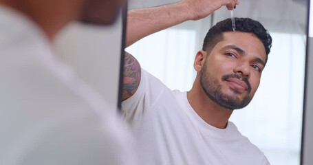 Handsome Asian man drops hair growth serum on his head while standing in front of a mirror.