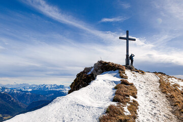 Ein Wanderer lehnt am Hochgern Gipfelkreuz mit beeindruckende Aussicht über die Chiemgauer Berge...