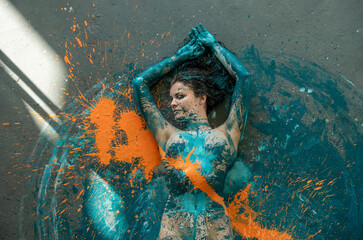 Upper body of young sexy naked woman in blue, orange and turquoise color painted, lies decorative, elegant on the Studio floor in shining light.
