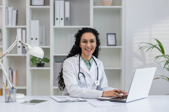 A cheerful Latino female doctor in a white coat smiles as she works on her laptop in a modern office setting.