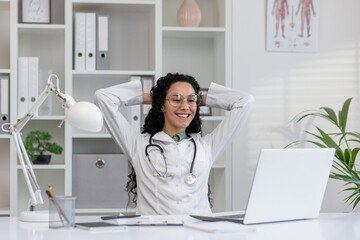 Obraz na płótnie Canvas Joyful Latina doctor in white coat taking a break at her office desk with laptop, feeling content and happy.
