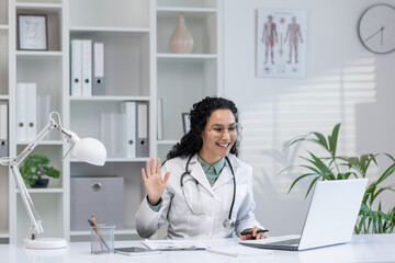 Friendly Hispanic female doctor in white coat waving during a video call consultation in a bright...