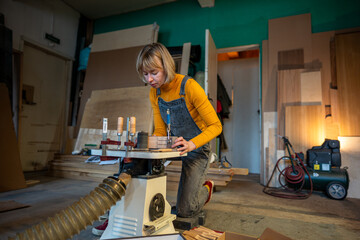Focused female joiner works in joinery workshop. Pedant woman sanding, turning wooden knife blanks on a lathe. Small family business, furniture workshop, production of customised wooden tableware. - obrazy, fototapety, plakaty