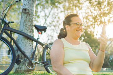 middle-aged latin woman sitting in a park next to her bike checking her cell phone happily.